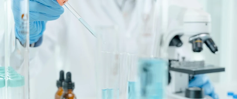 A person wearing a white lab coat and latex gloves holds a dropper of blue liquid above a test tube of the same fluid. There are other test tubes and a microscope on the table in front of the person. 